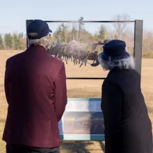Two people looking at a sign outdoors that displays a historical scene which appears on the winter-brown field in front of them.